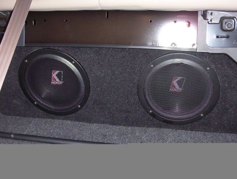2003 Ford Taurus - Two Kicker subwoofers with black mesh grilles mounted in a gray carpeted enclosure sitting in a sedan's trunk against the rear seat back. T…