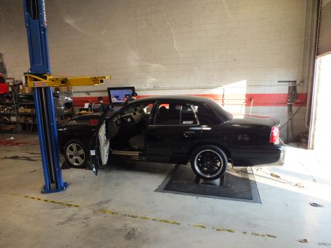 2003 Ford Crown Victoria - Black sedan with rear-quarter text staged on dyno rollers, door open against a painted block wall