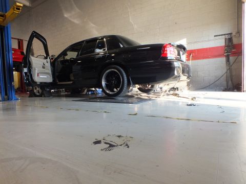 2003 Ford Crown Victoria - Low rear three-quarter of a black sedan on dyno rollers, door open, exhaust pipe trailing across floor