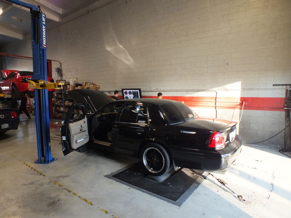 Black Ford sedan rear three-quarter on dyno, door open, red Mustang on lift behind