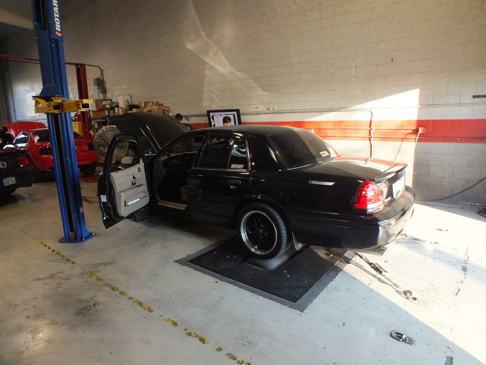 Black sedan rear three-quarter on dyno with open door, tuning monitor and lift in shop
