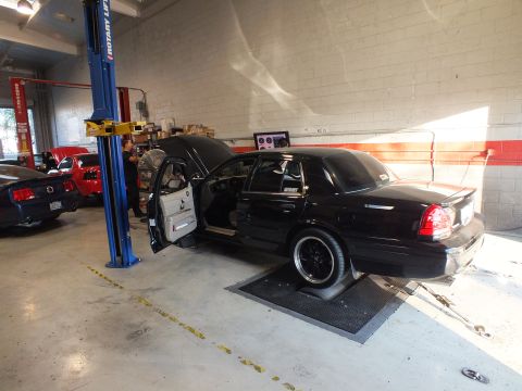 2003 Ford Crown Victoria - Black sedan on dyno rollers with door open; a blue Mustang and shop lifts fill the background