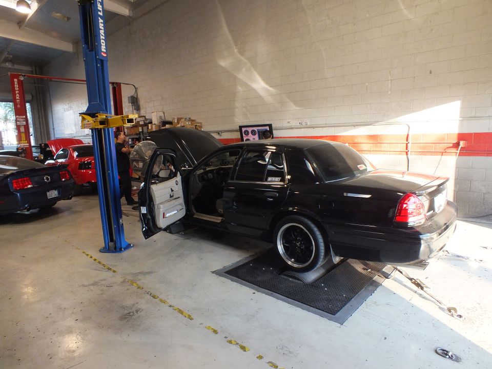Black sedan rear three-quarter on dyno, blue Mustang and lifts in shop background