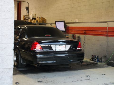 2003 Ford Crown Victoria - Black sedan strapped to a chassis dyno, exhaust tips visible, tuning monitor on the wall behind.