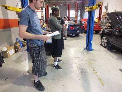 2003 Ford Crown Victoria - Man in gray tee reads a printout near another worker; lifts, a Mustang and dark sedan fill the shop.