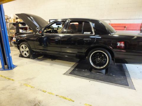 2003 Ford Crown Victoria - Driver-side view of a black sedan on the dyno, hood raised, black multi-spoke wheels fitted.
