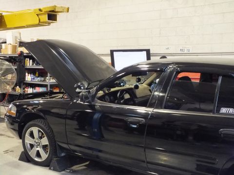 2003 Ford Crown Victoria - Open-hood black sedan tilted toward camera, monitor and supply shelves along the shop wall.
