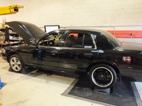 2003 Ford Crown Victoria - Side profile of a black sedan on dyno rollers, hood open, tuning screen mounted on the wall.