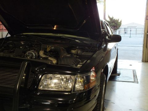 2003 Ford Crown Victoria - Tight front view of a black sedan, hood raised over the engine, one headlight lit, bay door behind.