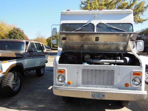 1979 Ford Bronco - A white delivery truck sits parked with its hood open, revealing the engine compartment, next to a brown and white SUV.