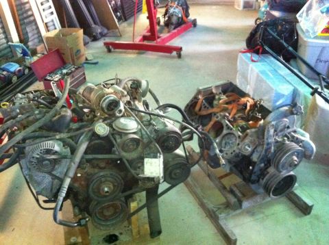 1986 Ford Bronco - Two engines are displayed on stands in a cluttered garage setting.