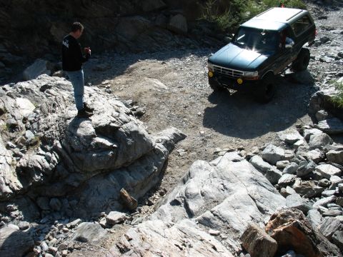 1994 Ford Bronco - A 1994 Ford Bronco tackles a challenging, rocky terrain on an off-road trail, with a person standing on a rock observing.