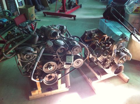 1986 Ford Bronco - Two V8 engines are displayed on wooden stands in a workshop setting, with tools and equipment visible in the background.
