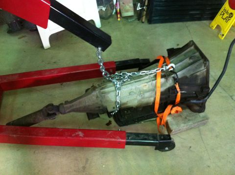 1986 Ford Bronco - A transmission is suspended by chains and straps from an engine hoist, ready for removal or installation.