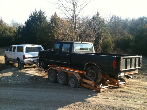 1986 Ford Bronco - A black Ford F-Series truck is being towed by a white Ford Bronco on a dirt road with trees in the background.
