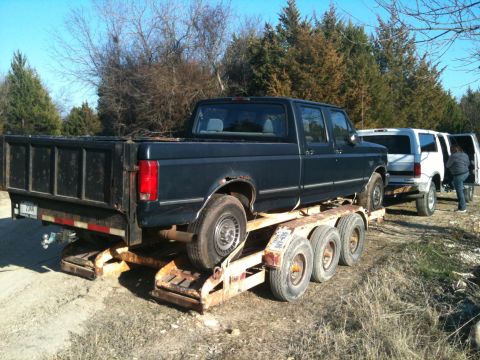 1986 Ford Bronco - A dark blue Ford F-Series pickup truck is loaded onto a trailer, with a white van behind it.