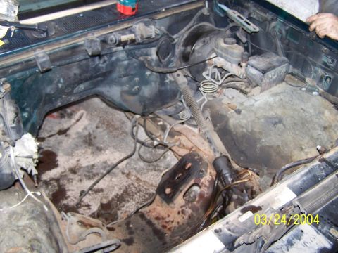 1986 Ford Bronco - The engine bay of a green vehicle shows significant rust and dirt, with visible brake components and steering column.