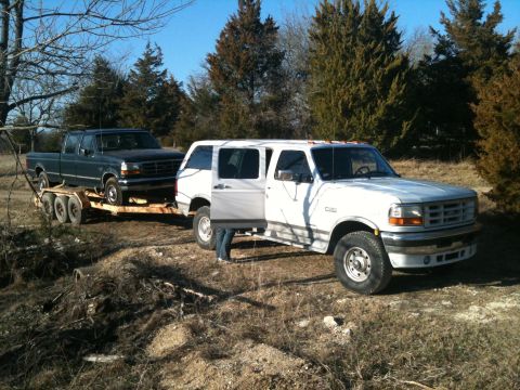 1986 Ford Bronco - A white 1995 Ford F-250 Super Duty tows a dark Ford F-150 on a trailer through a rural landscape.