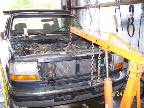 1986 Ford Bronco - The front of a dark-colored SUV is shown with its hood open and engine removed, supported by an engine hoist.