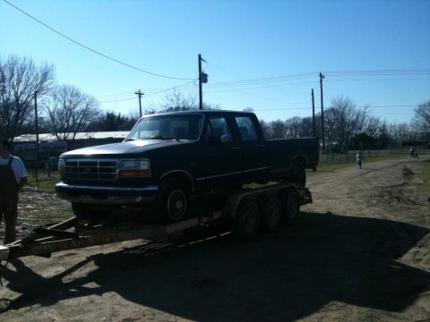 1986 Ford Bronco - A dark-colored pickup truck is loaded onto a triple-axle trailer, viewed from the front three-quarter angle.