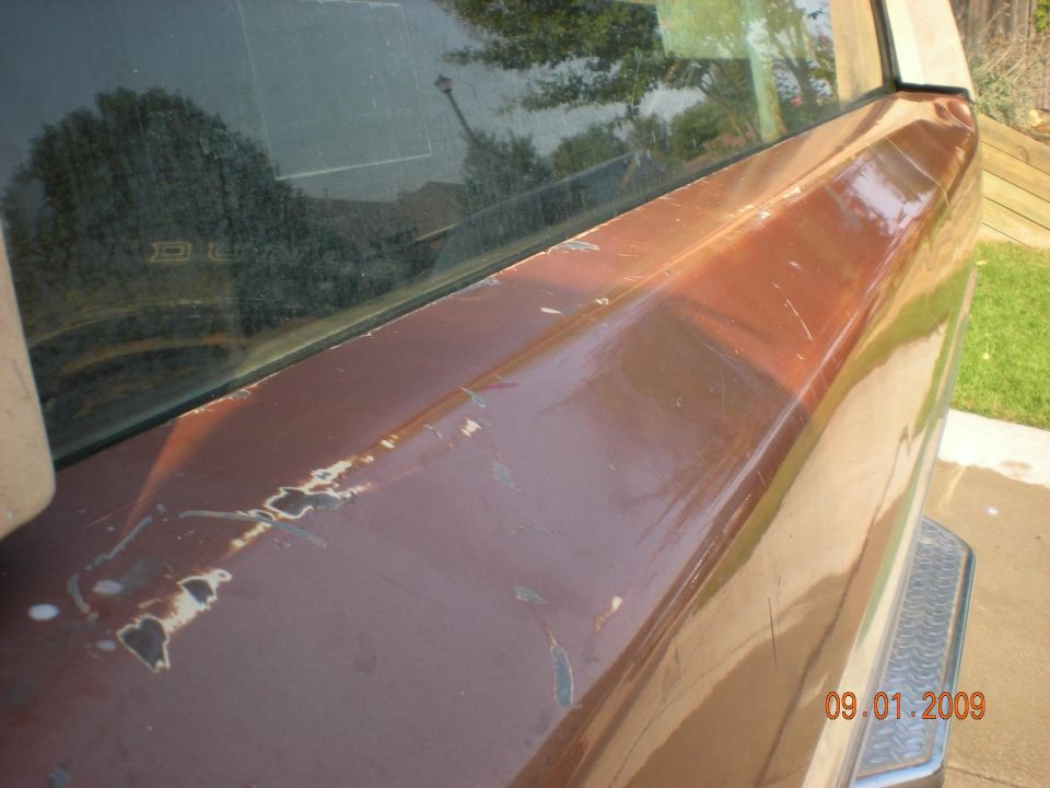 Brown SUV rear quarter panel with peeling paint and scratches, reflecting trees and sky