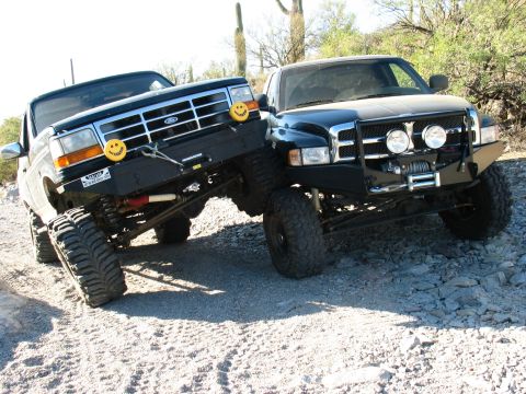1994 Ford Bronco - Two modified trucks, a Ford and a Dodge, are parked on a rocky, off-road trail with cacti in the background.