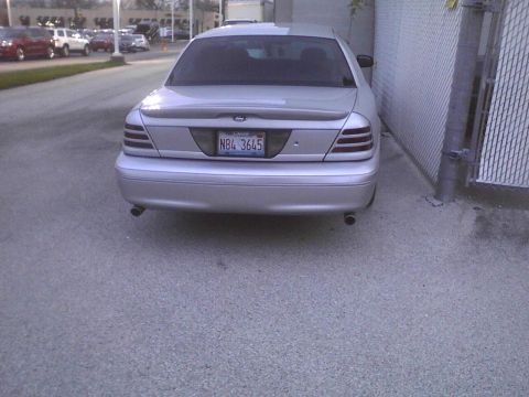 2003 Ford Crown Victoria - The rear of a silver 2003 Ford Crown Victoria sedan is visible, parked tightly beside a chain-link fence.
