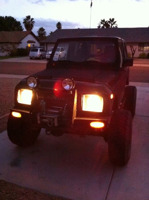1987 Suzuki Samurai - This Suzuki Samurai is parked in a residential driveway as the sun sets, with its headlights and marker lights illuminated.