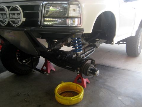 1996 Ford Bronco - Front suspension of a white SUV, possibly a 1992 Ford Bronco, undergoing maintenance with the front end raised on jack stands.