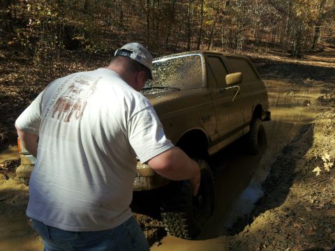 1986 Ford Bronco - A man inspects the wheel of a mud-covered 1986 Ford Bronco stuck in a muddy off-road trail.