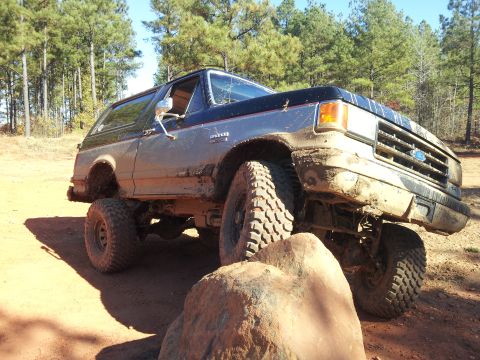 1986 Ford Bronco - This lifted Ford Bronco navigates a rocky trail, its tires caked in mud.