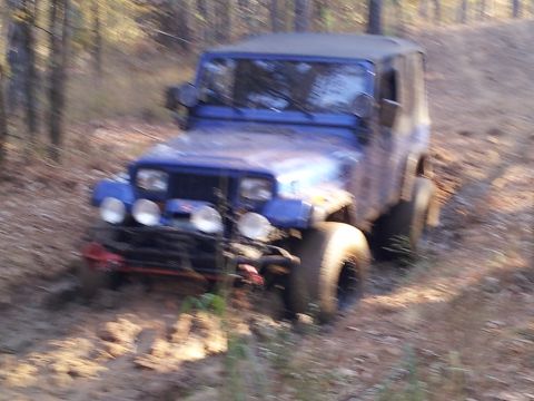 1986 Ford Bronco - A blue Jeep Wrangler navigates a muddy trail through a wooded area.