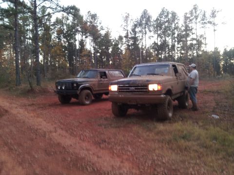 1986 Ford Bronco - Two Broncos are parked on a dirt road, one tan and one dark, with a man standing by the tan one.