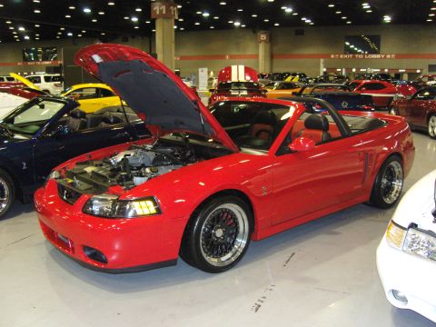 2007 Ford Mustang - Red convertible Mustang at a car show, hood open, with other vehicles in the background.