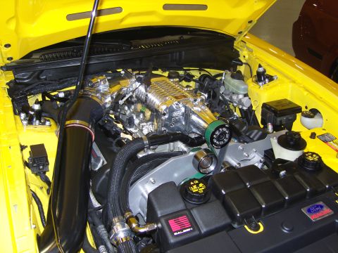 2007 Ford Mustang - Close-up of a supercharged engine bay in a yellow vehicle, a prominent chrome supercharger and a Ford badge.