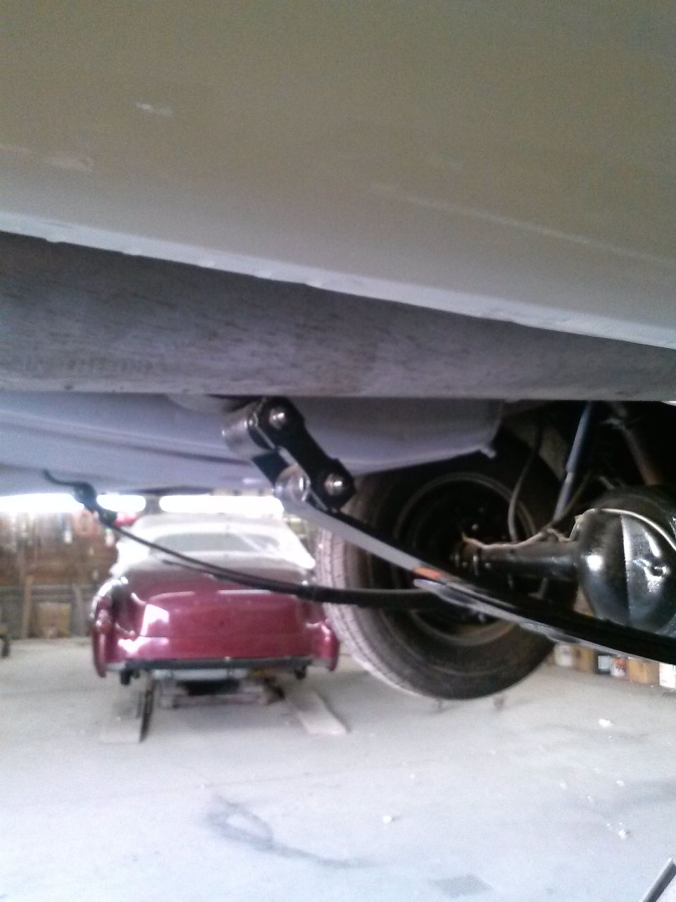 Rear suspension and leaf spring on a classic car, with a red coupe visible in the background