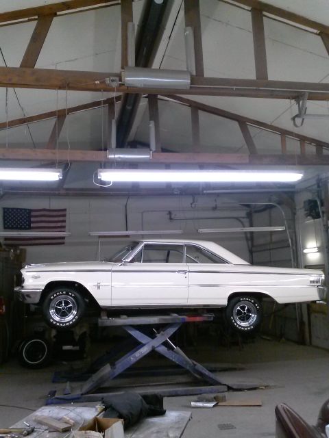 1960 Ford Falcon - A white 1963 Ford Galaxie coupe is elevated on a car lift inside a garage, with an American flag visible on the wall.
