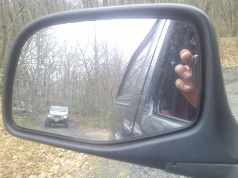 1995 Ford Bronco - A white Jeep Wrangler is visible in the side mirror of another vehicle on a dirt road surrounded by trees.