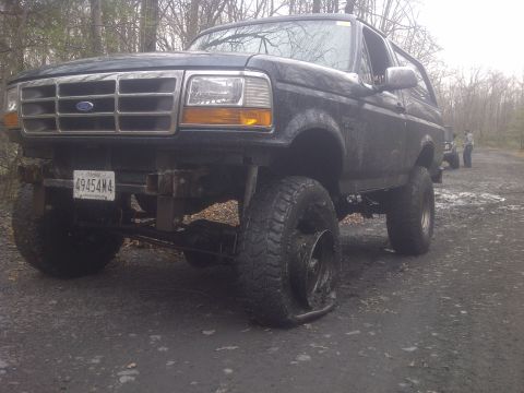1995 Ford Bronco - A black 1995 Ford Bronco sits with a flat tire on a muddy road, with other vehicles visible in the background.