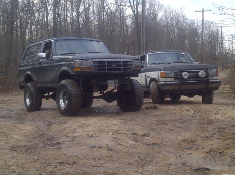 1995 Ford Bronco - Two off-road vehicles, a black Ford Bronco and a white pickup truck, are parked on a muddy trail surrounded by trees.
