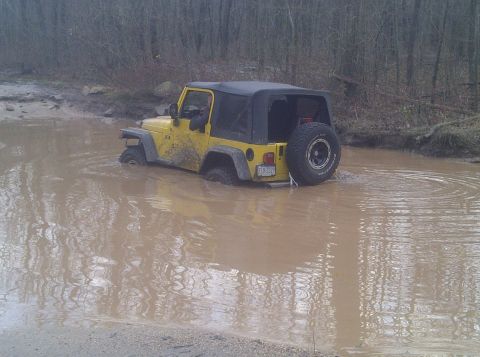 1995 Ford Bronco - A yellow Jeep Wrangler is stuck in a large mud puddle with trees in the background. The vehicle's tires are submerged in the murky water.