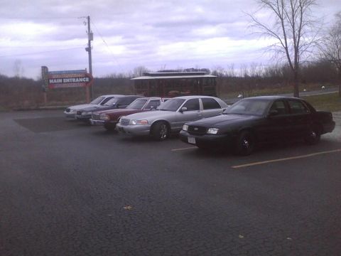 2003 Ford Crown Victoria - A line of sedans, including a black 2003 Ford Crown Victoria, are parked in a lot under an overcast sky.