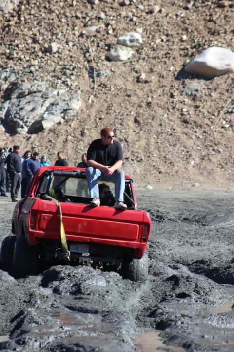 1978 Ford Bronco - A red pickup truck is stuck in deep mud with several people watching.