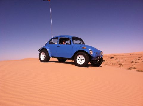 1970 Volkswagen Beetle - A blue 1970 Volkswagen Beetle Baja Bug navigates sandy terrain under a clear blue sky, a flag attached to its antenna.