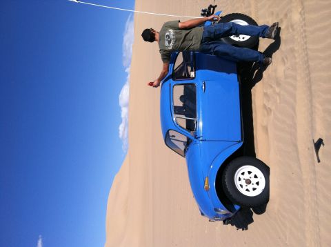 1970 Volkswagen Beetle - A man relaxes on a blue dune buggy parked on a sandy landscape under a clear blue sky.