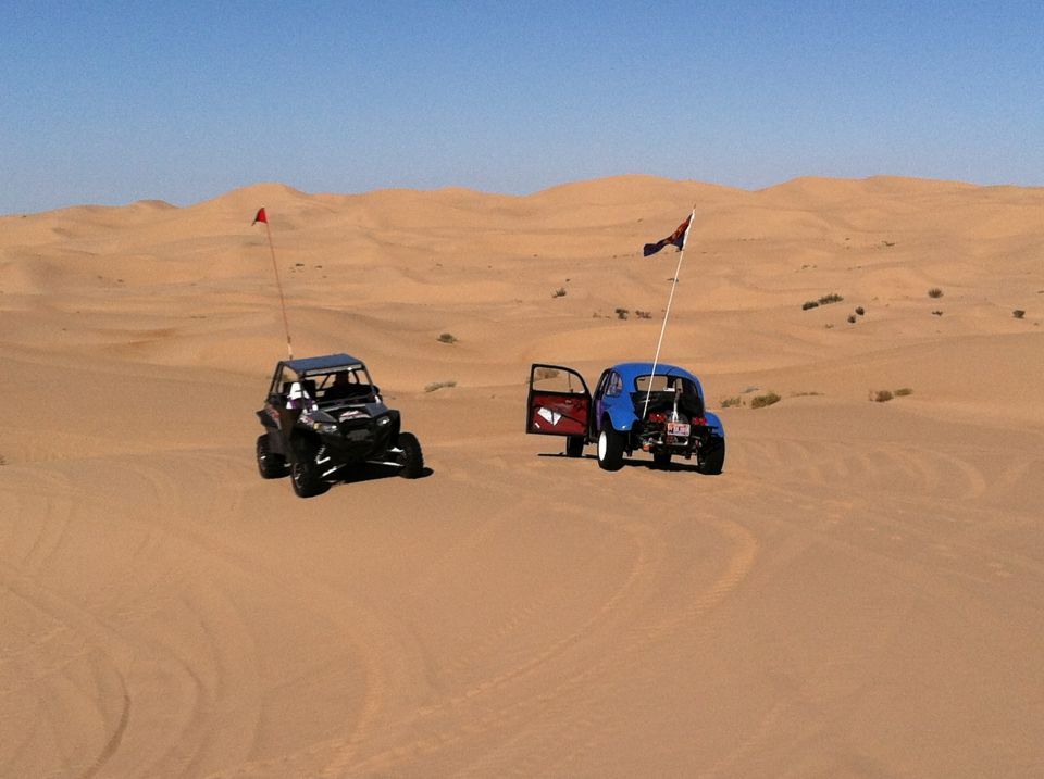 Blue 1970 Volkswagen Beetle with open door and black off-road vehicle in desert dunes