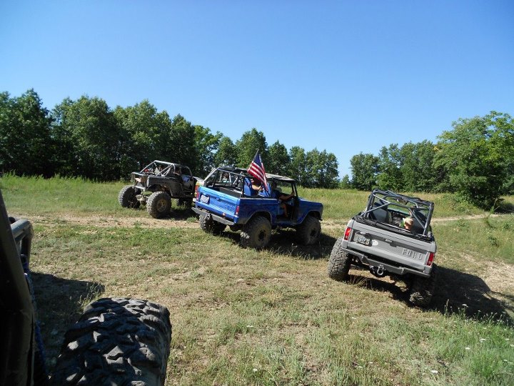 Blue Ford Bronco with American flag, gray Ford Bronco, and off-road buggy on a sunny day