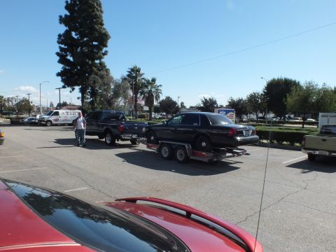 2003 Ford Crown Victoria - Black sedan strapped to a tandem-axle car trailer behind a dark pickup in a sunny lot.