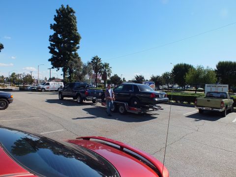 2003 Ford Crown Victoria - Man stands beside a black sedan loaded on a trailer hitched to a pickup, lot and palms behind.