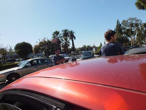 2003 Ford Crown Victoria - View over a red car roof of two men chatting; a tan Crown Victoria and palms fill the lot.